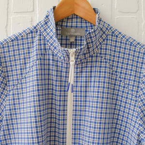 May include: A blue and white checkered zip-up jacket hanging on a wooden hanger. The jacket has a high collar and a white zipper. The fabric is a classic gingham pattern, with a small tag visible at the collar.