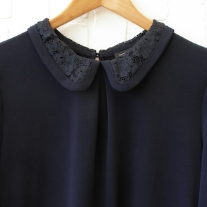 May include: A navy blue blouse with a Peter Pan collar. The collar is trimmed with black lace. The blouse has long sleeves and is hanging on a wooden hanger.