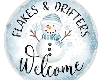 May include: Circular winter sign featuring a snowman illustration. The snowman has a blue and white striped hat and scarf, a carrot nose, and twig arms. The sign displays the text "Flakes & Drifters Welcome" in black script on a snowy blue backdrop.