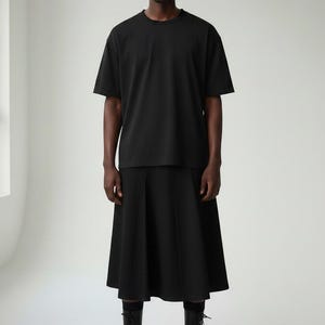 Op de afbeelding: Een persoon draagt een zwart T-shirt met ronde hals, een zwarte geplooide rok, zwarte laarzen en een zwarte zonnebril. De outfit is volledig zwart, wat een minimalistische en moderne esthetiek creëert. De laarzen hebben een vetersluiting.