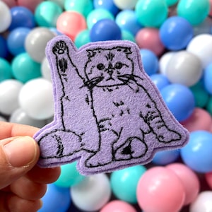 May include: A purple felt patch with an embroidered image of a cat waving its paw. The cat is black and white and has a surprised expression.