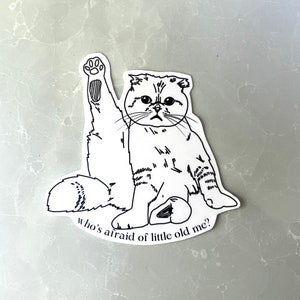 May include: A black and white sticker featuring a Scottish Fold cat with one paw raised. The cat has a grumpy expression and the text "who's afraid of little old me?" is below the cat.