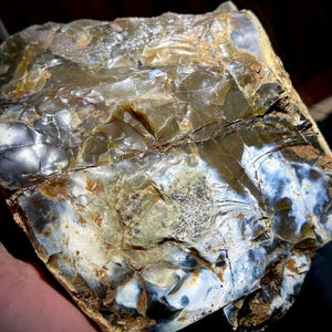 May include: A rough, irregularly shaped piece of agate with a mix of brown, white, and blue colors. The agate has a layered appearance with a shiny, iridescent surface.