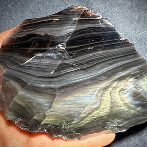May include: A large, irregularly shaped piece of obsidian with a layered, banded appearance. The stone displays a range of colors, including black, gray, and hints of iridescent blues and greens. The surface is smooth and polished, with visible striations.