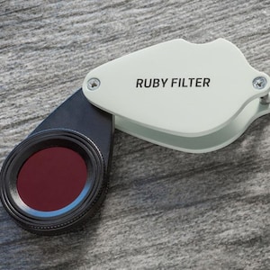May include: A white and black magnifying glass with a red filter. The magnifying glass has the words "RUBY FILTER" printed on the side.