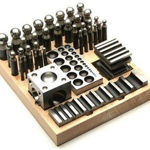 May include: A wooden box filled with metal tools for jewelry making. The box contains a variety of round and square punches, a set of metal rings, and a set of metal stamps.