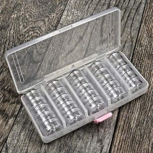 May include: A clear plastic storage container with a hinged lid, divided into five compartments. Each compartment holds several small, clear, screw-top jars. The container is closed with a pink clasp. The background is a wooden surface.