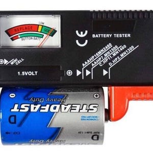 May include: A black battery tester with a red and black probe. The tester has a digital display and a dial with a needle. The tester is labeled "BATTERY TESTER" and lists the battery sizes it can test: AAA, AA, C, D, and HP2. The tester is testing a blue and white battery labeled "STEADFAST Heavy Duty".