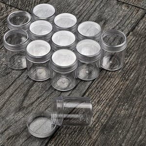 May include: A set of 12 clear plastic jars with lids. The jars are arranged in a circle on a wooden surface.