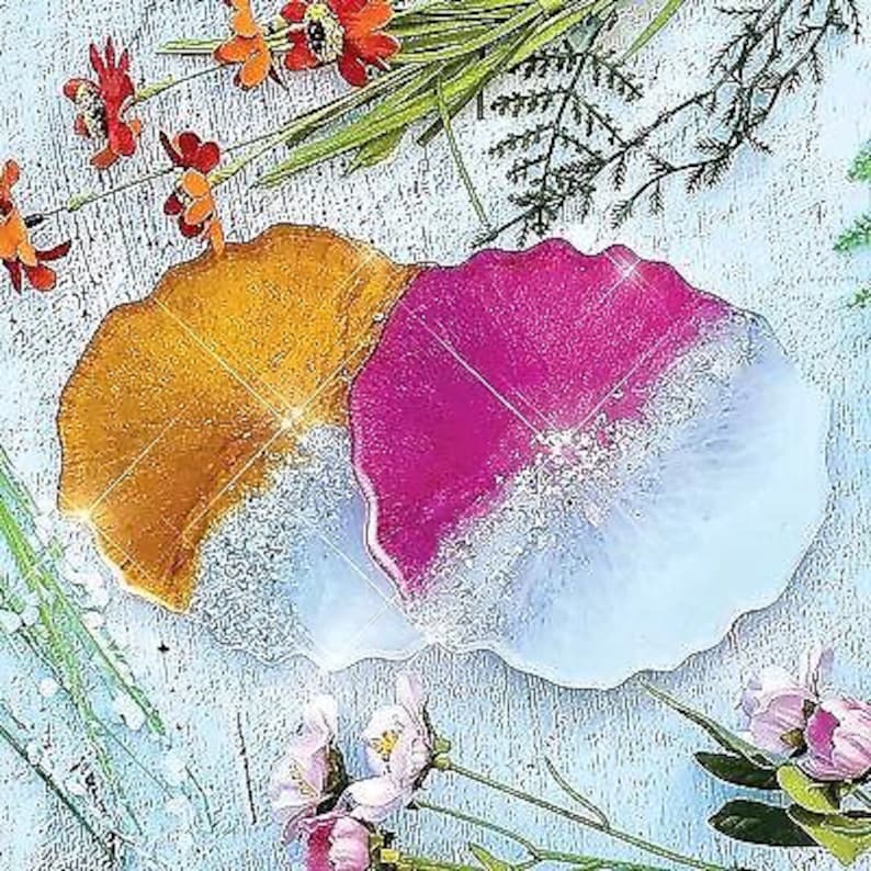 K&ouml;nnte beinhalten: Zwei Untersetzer aus Harz mit einem einzigartigen, flie&szlig;enden Design. Ein Untersetzer ist leuchtend pink und wei&szlig;, w&auml;hrend der andere warm gold und wei&szlig; ist.