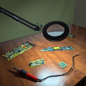 May include: A black articulated magnifying lamp illuminates circuit boards, a soldering iron, and a CPU on a wooden surface. The lamp has a large circular lens. Various electronic components are scattered across the table.
