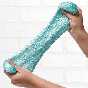 May include: A light blue slime with a glittery finish and small white beads throughout. The slime is being stretched and pulled apart by two hands.