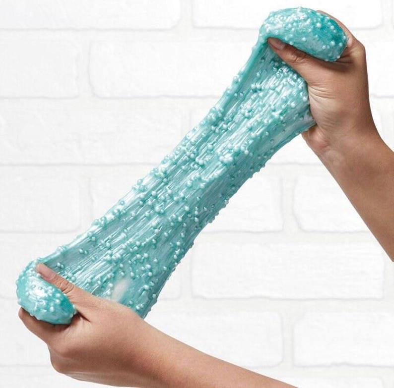 May include: A light blue slime with a glittery finish and small white beads throughout. The slime is being stretched and pulled apart by two hands.