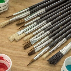 May include: A set of paintbrushes with wooden handles and silver metal ferrules. The brushes are arranged in a fan shape on a wooden surface. There are also several small containers of paint in the background.
