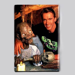 Op de afbeelding: Een rechthoekige magneet met een scène van een man in een zwart T-shirt met de tekst TALES FROM THE CRYPT in groene letters. Naast hem staat een skeletachtige figuur met blauwe ogen die een menselijke schedel bij een brandende kaars houdt.