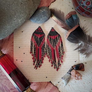 Può includere: Un paio di lunghi orecchini a perline con uno schema di colori nero e rosso. Gli orecchini sono realizzati con piccole perline rotonde e hanno un design a frangia.