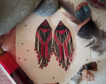 Puede incluir: Un par de pendientes largos de cuentas con un esquema de color negro y rojo. Los pendientes están hechos con cuentas pequeñas y redondas y tienen un diseño de flecos.