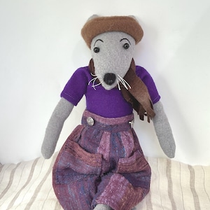May include: A handmade stuffed animal mouse wearing a brown hat, scarf, and purple shirt. The mouse has gray arms and a gray face with a black nose and white whiskers. It is wearing purple and brown patterned pants with silver buttons.