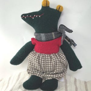 May include: A handmade stuffed animal frog with a dark green body, red shirt, and houndstooth patterned shorts. The frog has a gray scarf and yellow accents on its head. The frog is sitting on a striped fabric.