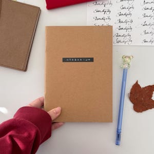 Op de afbeelding: Een bruin notitieboek met het woord "HERBARIUM" gedrukt op een zwart rechthoek. Een blauwe pen met een berenvormige topper en een bruin blad liggen naast het notitieboek. Een rode trui is zichtbaar in de hoek.