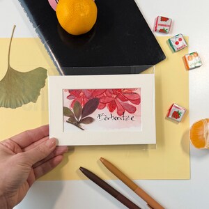 Puede incluir: Una impresión artística botánica enmarcada con hojas rojas y verdes y la palabra "Berberitze". La impresión está sobre un fondo amarillo con un cuaderno negro, caramelos, bolígrafos y una mandarina.
