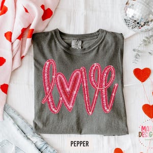May include: A gray t-shirt with the word "LOVE" in pink glitter lettering. The shirt is surrounded by a pink sweater with red hearts, light blue jeans, and red heart decorations. The word "PEPPER" is printed below the shirt.