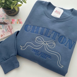 Könnte beinhalten: Ein blaues Sweatshirt mit dem Wort "CHILTON" in dunklerem Blau gestickt, zusammen mit einer Schleife und den Worten "Prep School Est 1863". Ein Notizbuch, ein Stift und eine kleine Pflanze befinden sich im Hintergrund.
