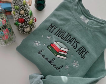 Puede incluir: Una sudadera de color verde azulado con el texto "MY HOLIDAYS ARE booked" bordado en la parte delantera. El texto está rodeado de pequeños diseños de copos de nieve.
