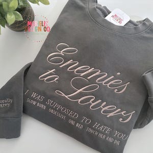 May include: A gray sweatshirt with the embroidered text "Enemies to Lovers" in a cursive font. Below the main text are additional embroidered phrases. The left sleeve cuff reads "morally grey". The sweatshirt is a casual apparel item.