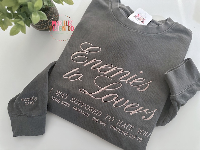 May include: A gray sweatshirt with the embroidered text "Enemies to Lovers" in a cursive font. Below the main text are additional embroidered phrases. The left sleeve cuff reads "morally grey". The sweatshirt is a casual apparel item.