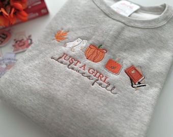 Könnte beinhalten: Ein meliertes graues Sweatshirt mit gestickten Herbstmotiven. Das Design umfasst einen Kürbis, Socken, ein Blatt, eine Parfümflasche und ein Buch. Der Text "Just a girl who loves fall" ist in Schreibschrift gestickt.
