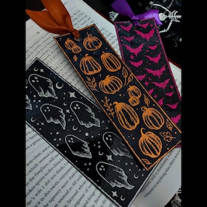 Può includere: Tre segnalibri a tema Halloween con disegni arancioni, rosa e bianchi. Un segnalibro nero presenta fantasmi, stelle e lune bianche. Un altro segnalibro nero ha zucche arancioni. Il terzo è rosa con sagome di pipistrelli.