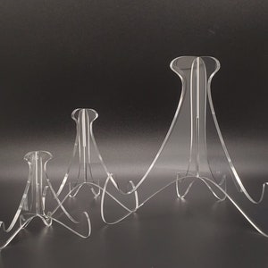 Könnte beinhalten: Drei transparente Acryl-Ausstellungsständer in verschiedenen Größen. Jeder Ständer hat eine gebogene Basis und ein hohes, offenes Design auf der Rückseite. Sie sind für die Präsentation von Gegenständen wie Tellern oder Kunstwerken konzipiert.