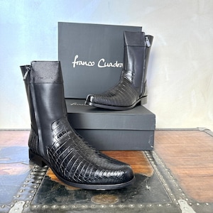 Peut inclure: Une paire de bottes en cuir noir à motif crocodile. Les bottes ont une fermeture éclair sur le côté et une pointe pointue. Les bottes sont posées sur une surface en bois avec une boîte derrière elles. La boîte est noire et porte l'inscription "Franco Cuadra".