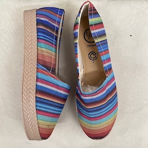May include: A pair of blue and red striped canvas espadrille shoes with a platform wedge sole. The shoes have a woven fabric upper and a comfortable, casual style.
