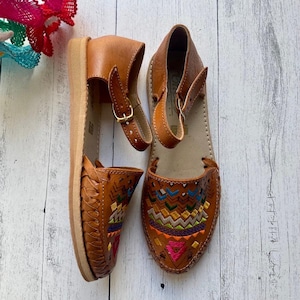 May include: A pair of brown leather sandals with colorful embroidered designs on the toe. The sandals have an ankle strap with a buckle closure and a woven design on the front. The sandals are displayed on a white wooden surface.