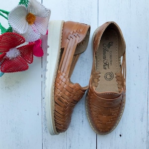 May include: A pair of brown leather sandals with a woven design. The sandals have a white platform sole and are made by the Amerí Hu Collection.