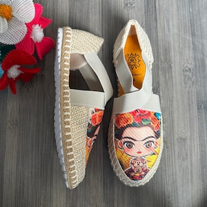 Può includere: Ballerine espadrillas con design Frida Kahlo. Le scarpe presentano un tessuto beige intrecciato, un cinturino elastico beige e una suola bianca. Il design Frida Kahlo include un copricapo floreale e un cagnolino. Taglia 4.
