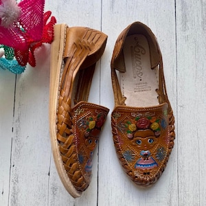May include: A pair of brown leather sandals with colorful embroidery. The sandals have a woven design and feature a floral pattern with a small figure in the center. The sandals are labeled "Lucas" on the inside.