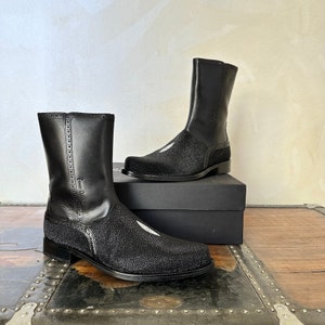 May include: A pair of black leather boots with a pointed toe and a textured design. The boots have a tall shaft and a zip on the side. The boots are sitting on a wooden surface.