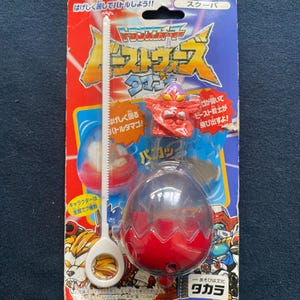 May include: Vintage Transformers toy package featuring a red and blue egg-shaped toy with a white handle and a small red figure. The packaging has Japanese text and colorful graphics.