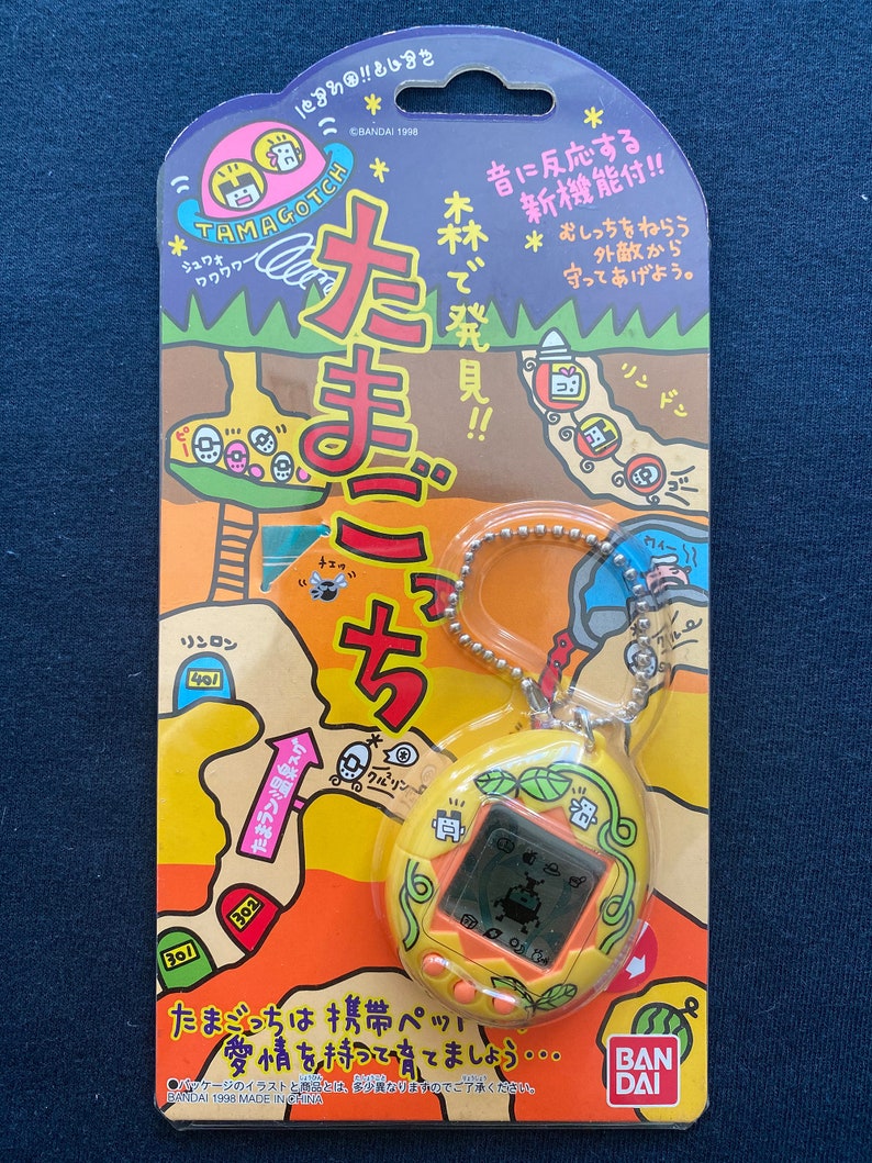 May include: A yellow Tamagotchi keychain with a green leaf design. The screen shows a small, black creature with a white outline. The packaging features a cartoon illustration of a Tamagotchi character and Japanese text.