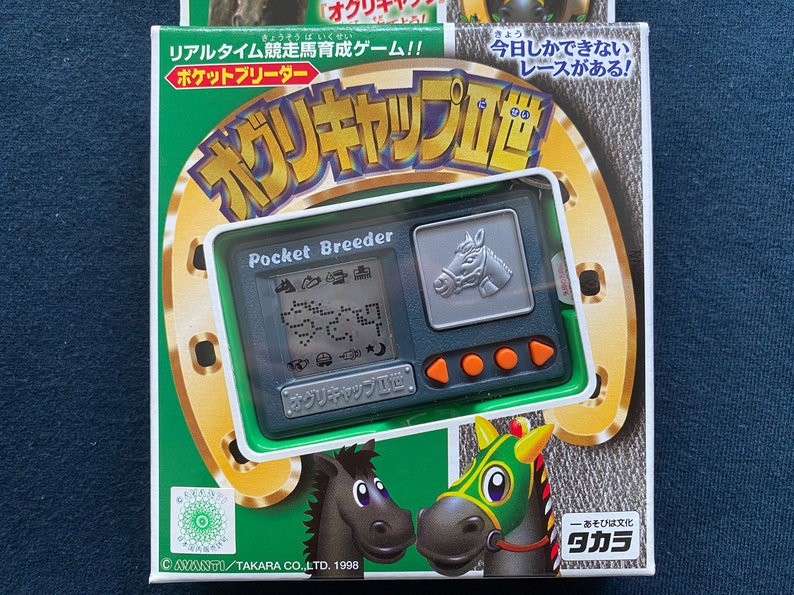 May include: A green and gold box with a silver and green handheld electronic game. The game is called "Pocket Breeder" and features a horse on the screen. The box also has the text "Oguri Cap II" and "Takara" on it.