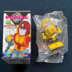 Op de afbeelding: Een rood en geel Transformers-actiefiguur in een plastic zak. De figuur is gelabeld met "MYCLONE" en "AS-4". De figuur is verpakt in een kartonnen doos met een rood en geel Transformers-actiefiguur op de voorkant.
