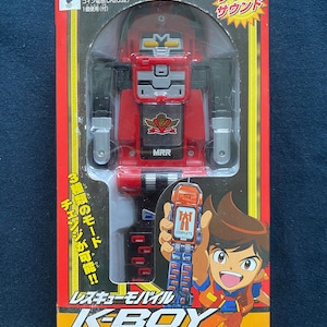 Puede incluir: Un robot de juguete rojo y negro con un teléfono móvil sujeto a su pecho. El robot tiene una placa de pecho plateada con un logotipo que dice "MRR". El juguete está empaquetado en una caja con un niño de dibujos animados en la parte delantera. La caja dice "Rescue Mobile K-BOY" y "K-BOY" en letras grandes. La caja también dice "Luz y sonido" y "¡3 modos de cambio posibles!" en japonés.