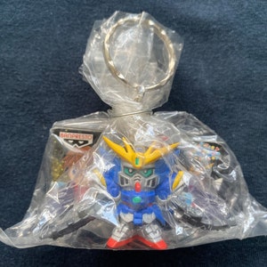 May include: A blue and yellow plastic keychain featuring a robot character with a silver key ring. The keychain is packaged in a clear plastic bag with a silver key ring. The packaging also includes a small card with the text "BANPRESTO" printed on it.