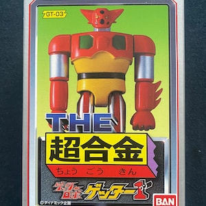 Puede incluir: Un juguete robot rojo y amarillo con el texto "THE 超合金 ちょうごう きん" y "ゲッター1" en la caja. La caja es blanca con una franja azul y roja y el texto "BAN DAI" en la esquina inferior derecha.