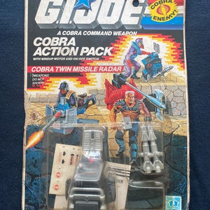 May include: A vintage G.I. Joe Cobra Twin Missile Radar action figure pack. The packaging features a red, white, and black color scheme with the Cobra logo and the words "Cobra Enemy!" The figure is a silver and black missile launcher with a red button. The packaging also includes a warning label that says "Weapons Do Not Shoot".