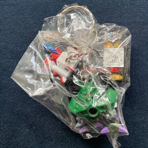 May include: A green and purple robot keychain with a red eye and a silver ring. The keychain is packaged in a clear plastic bag with a white and red spaceship keychain.