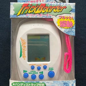 Pode incluir: Jogo portátil Trick Boarder branco na embalagem original. O jogo apresenta um ecrã pequeno, botões azuis e um cordão rosa. A embalagem tem texto japonês e um gráfico de um snowboarder.